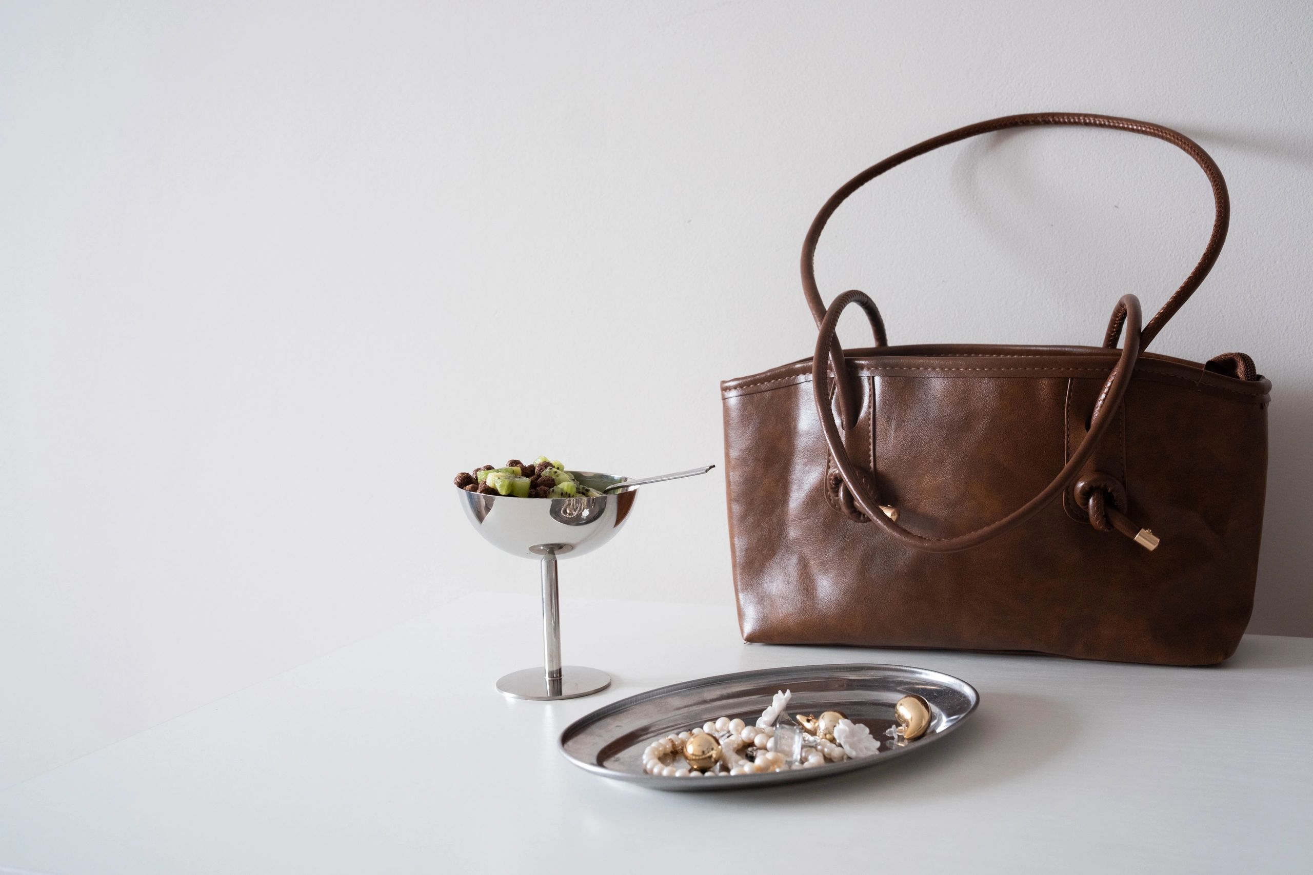 A stylish brown leather tote bag sits beside a metallic dessert bowl with fruit and a silver tray with jewelry on a clean white background minimalist chic composition.